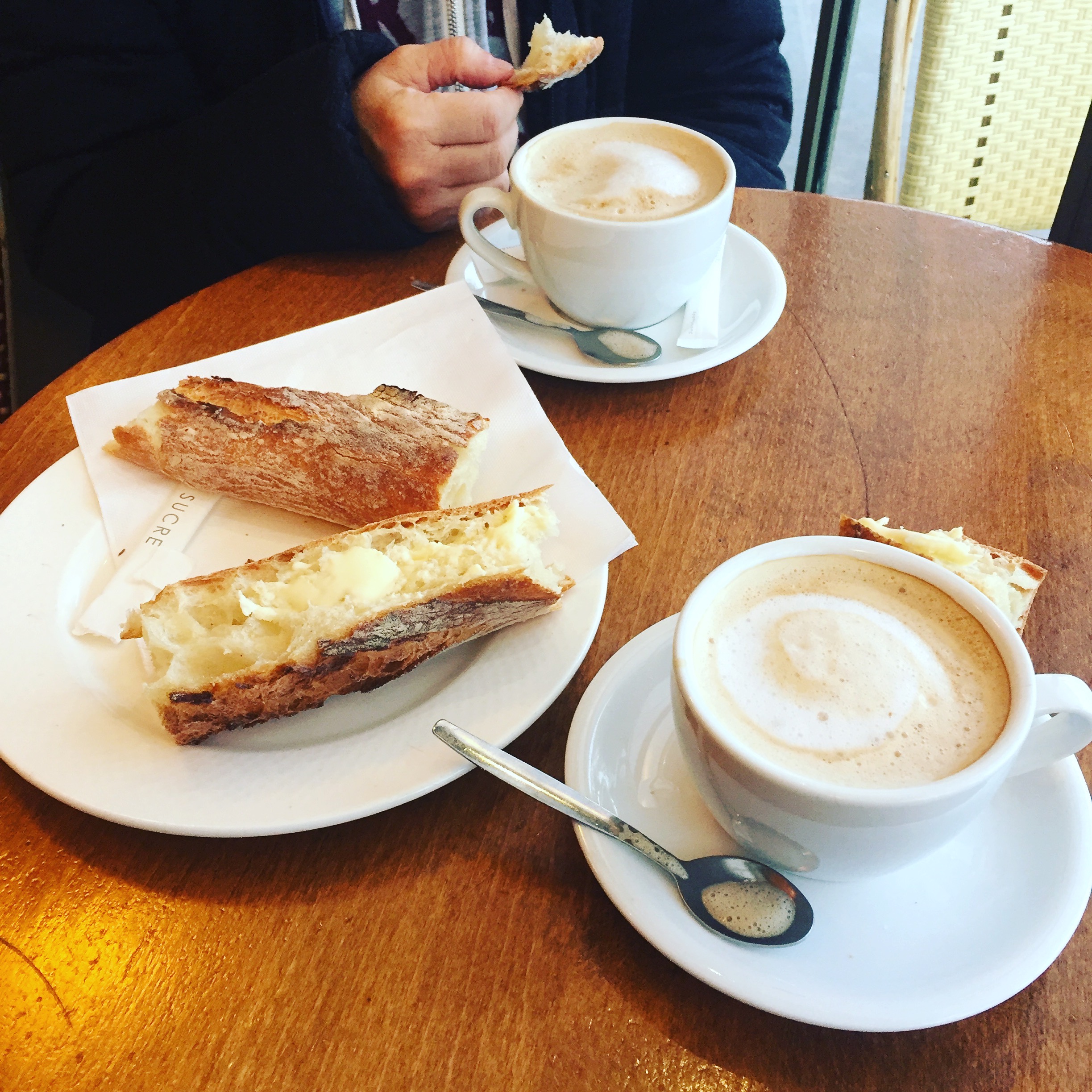 A buttered baguette and cappucinos for two on a table, photographed by Jeff Kraft